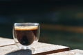 Small transparent double wall glass cup of fresh brewed coffee placed on a natural rough wooden surface in front of a decorative