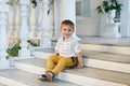 Small very cute, charming boy in yellow trousers sits on the sta