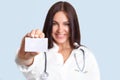 Small white blank card with copy space in hand`s of pretty nurse. Lovely brunette professional cheerful female family doctor with Royalty Free Stock Photo