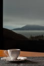 Small white coffee set with a biscuit on the side, on a wooden table. In the background a sea and a mountain landscape on a cloudy Royalty Free Stock Photo