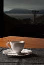 Small white coffee set with a biscuit on the side, on a wooden table. In the background a sea and a mountain landscape on a cloudy