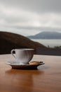 Small white coffee set with a biscuit on the side, on a wooden table. In the background a sea and a mountain landscape on a cloudy Royalty Free Stock Photo