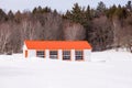 Small white hen house with orange roof and window trim in snowy field seen during a sunny early spring afternoon