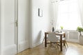 A small wooden dining table with chairs in a sunny room interior with white walls, double door and a window. Real photo.