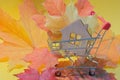 A small wooden house in a shopping cart against a background of autumn yellow-red maple leaves. Idea - autumn discounts, sale,