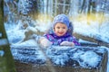 Small 4 yaers old girl in snowy winter forest plays outdoor. Child in colorful clothes smile and look away. Wooden trunk in front
