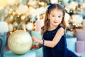 Small 4 years old beautiful girl in black fashion dress playing with big gold ball near christmas tree with christmas light and Royalty Free Stock Photo