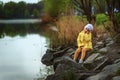 Small 5 years old girl in yellow coat and white hat sits on the river bank in spring time. Child on rock near water. Thinking Royalty Free Stock Photo