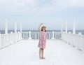Small 8 years old pretty cheerful girl in a dress with a floral print and in yellow sunglasses stands on a wooden white pier