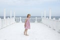 Small 8 years old pretty cheerful girl in a dress with a floral print and in yellow sunglasses stands on a wooden white pier