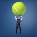 A small young muscular man tries to hold a giant yellow tennis ball above his head.