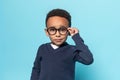 Smart african american little boy in school uniform touching glasses and looking at camera, standing on blue background
