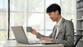 A smart Asian male boss reading business documents and sipping coffee at his desk Royalty Free Stock Photo