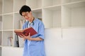Smart Asian male medical student wearing glasses and reading a book while standing in library Royalty Free Stock Photo