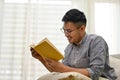 Smart Asian man wearing glasses reading a book on sofa in his living room. side view Royalty Free Stock Photo