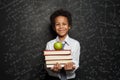 Smart black child student smiling and holding books and green apple against blackboard background with science and maths formulas