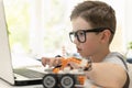 A smart boy builds a robot car using a laptop near the window at home. The child is learning coding and programming