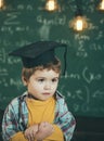 Smart child in graduate cap on serious face, shy, holds hands crossed. Kid, preschooler or first former, chalkboard on