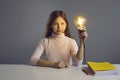 Portrait of serious school girl sitting at desk with books and holding glowing light bulb