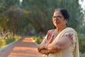 Smart and confident senior north Indian woman standing, thinking and looking away with hands crossed / folded in a park wearing an