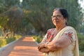 Smart and confident senior north Indian woman standing, thinking and looking away with hands crossed / folded in a park wearing an