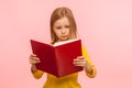 Smart cute little girl reading big book with serious attentive expression, learning homework