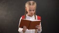 Smart female kid reading book, blackboard on background, thirst for knowledge