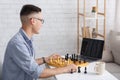 Smart guy and hobby in free time on self-isolation. Happy chess player in glasses with chess board looking at laptop