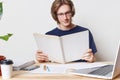 Smart hard working stylish male student wears spectacles, has attentive gaze in book, reads scientific literature before writing a Royalty Free Stock Photo