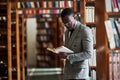 Smart looking african american businessman in glasses standing and reading a book in library