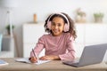 Smart teen girl in wireless headset having online lesson, using laptop and taking notes in her exercise book Royalty Free Stock Photo