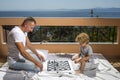 Smart toddler concept Dad with child play intellectual game Father and little son play chess on balcony nature and sea