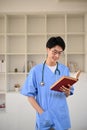 Smart young Asian male medical student in a uniform reading a book while standing in library Royalty Free Stock Photo