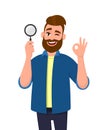 Smart young man holding magnifying glass and gesturing okay/OK sign while winking eye. Deal, good, agree, search, find, discovery.