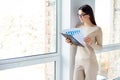 Smart young woman in glasses standing near large window and looking at the graphs and diagrams