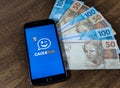 Smartphone with app (CAIXA TEM) on the screen with Brazilian real notes.