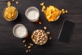 Smartphone on dark wooden table, nearby are nachos, chips and pistachios in glass plates and two glasses of beer