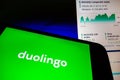 Smartphone with Duolingo language app logo on screen and blurred background with Nasdaq stock index chart. Duolingo will go public