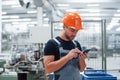 Smartphone in hands. Industrial worker indoors in factory. Young technician with orange hard hat