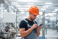 Smartphone in hands. Industrial worker indoors in factory. Young technician with orange hard hat