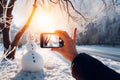 A smartphone in the hands of a tourist taking a photo of a friendly snowman in a city park on a frosty winter day