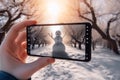 A smartphone in the hands of a tourist taking a photo of a friendly snowman in a city park on a frosty winter day