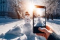 A smartphone in the hands of a tourist taking a photo of a friendly snowman in a city park on a frosty winter day Royalty Free Stock Photo