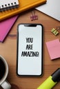 Smartphone with phrase You Are Amazing on screen and stationery against wooden background, flat lay