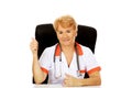 Smile elderly female doctor or nurse sitting behind the desk and shows thumb up Royalty Free Stock Photo