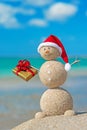 Smiley sandy snowman at beach in christmas hat with golden gift.