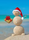 Smiley sandy snowman at beach in christmas hat with golden gift.