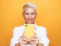 Smiley senior woman is holding a new smartphone in her hand over yellow background