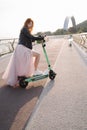 Beautiful happy young ginger girl using electric scooter in fashinable long pink dress black lether jacket