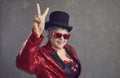 Happy senior woman in sequin outfit, top hat and cool glasses doing V sign gesture